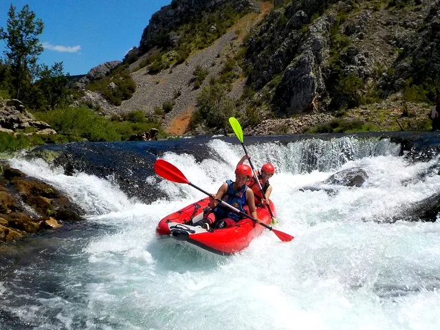 Deux personnes dans un kayak rouge pagayent dans de petits rapides par une journée ensoleillée.