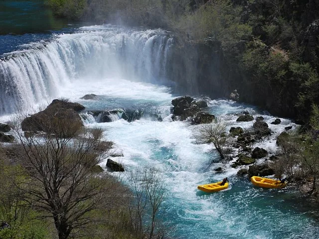 Deux radeaux jaunes naviguent dans les rapides sous une grande cascade, dans une gorge rocheuse.