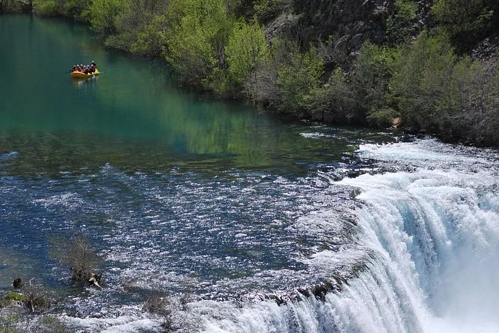 Ein großer Wasserfall stürzt über Felsen in ein klares Becken, in der Ferne ein Raft.